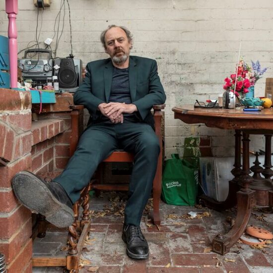 Man sitting in a chair wearing a green suit against a brick wall next to a table with flowers and a radio | Dan Tuffy Song of the Day Feature | Eat Sleep Breathe Music