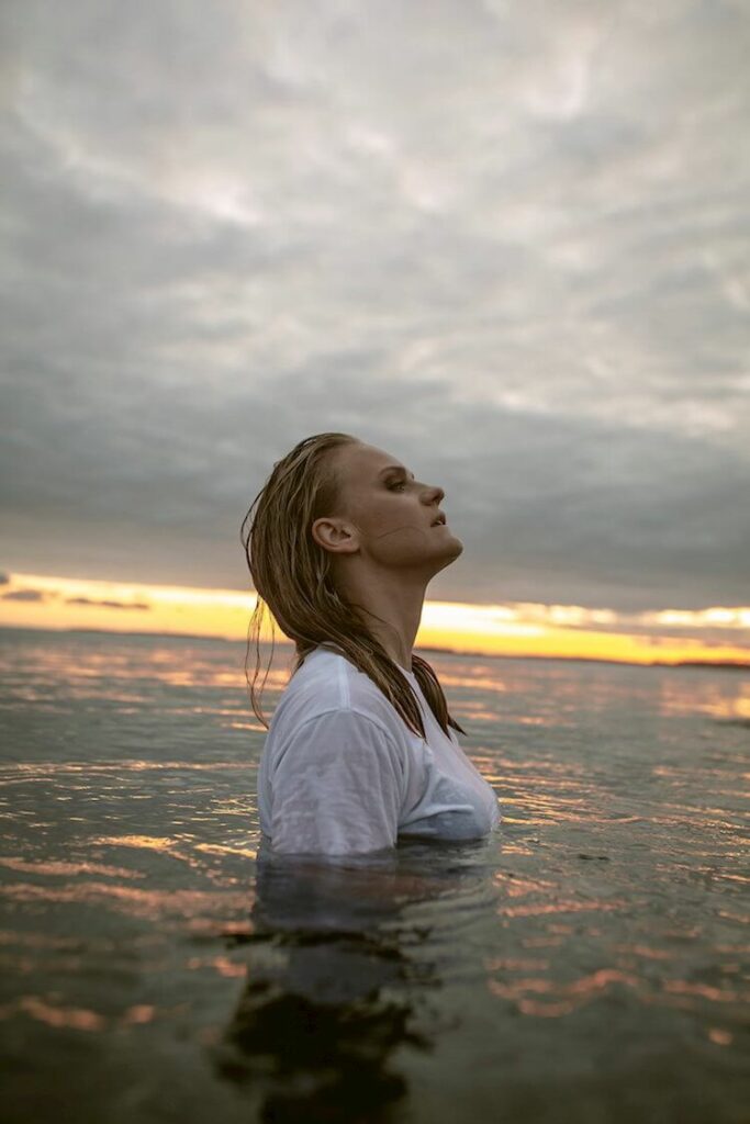 White Blonde Woman Standing in Water with White Tee shirt and and head thrown back against a dark sky and sunset Anni Pohto DEEP Song of the Day | Eat Sleep Breathe Music