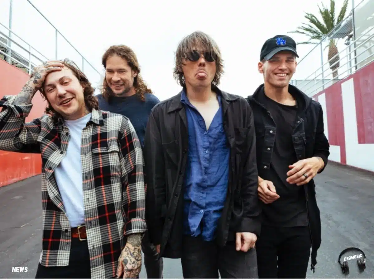 A candid photo of the four members of My Chemical Romance standing outside near a red and white wall with a chain-link fence and palm tree in the background. They are smiling and relaxed—Frank Iero wears a plaid flannel, Ray Toro is in a sweater, Gerard Way sticks out his tongue while wearing sunglasses and a dark jacket, and Mikey Way wears a black cap and hoodie while smiling broadly. | Eat Sleep Breathe Music