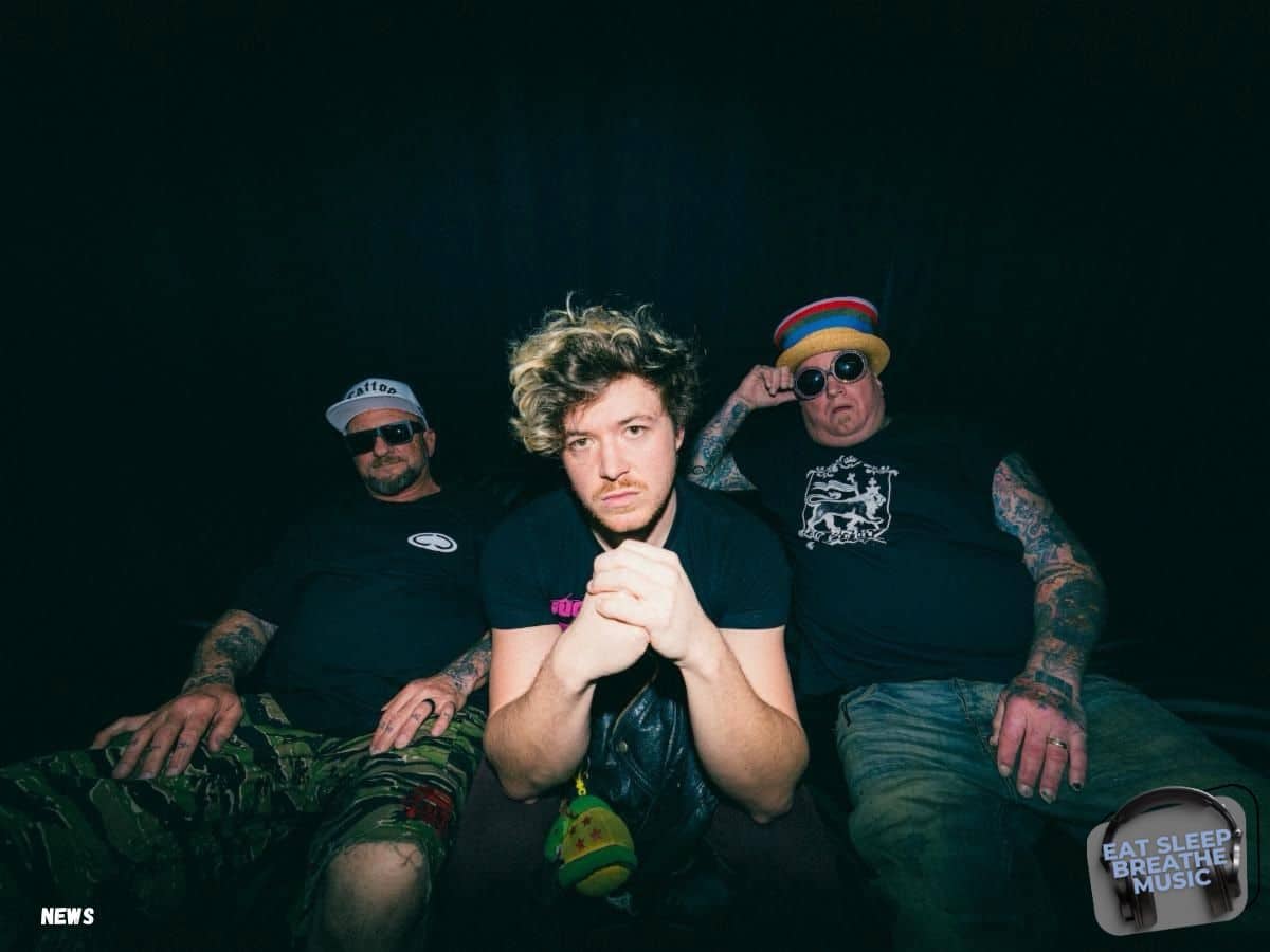 The three members of Sublime pose for a promotional photo against a dark backdrop. Jakob Nowell sits in the center with a serious expression, leaning slightly forward with hands clasped. Bud Gaugh sits to the right wearing a colorful beanie and sunglasses, and Eric Wilson sits to the left in a black shirt and camouflage pants, also wearing sunglasses and a hat. The image has a gritty, backstage vibe with a logo reading "Eat Sleep Breathe Music" in the corner. | Eat Sleep Breathe Music