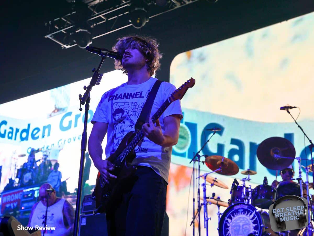 Jakob Nowell performing live with Sublime, playing guitar and singing during a concert in front of a bright screen displaying ‘Garden Grove."